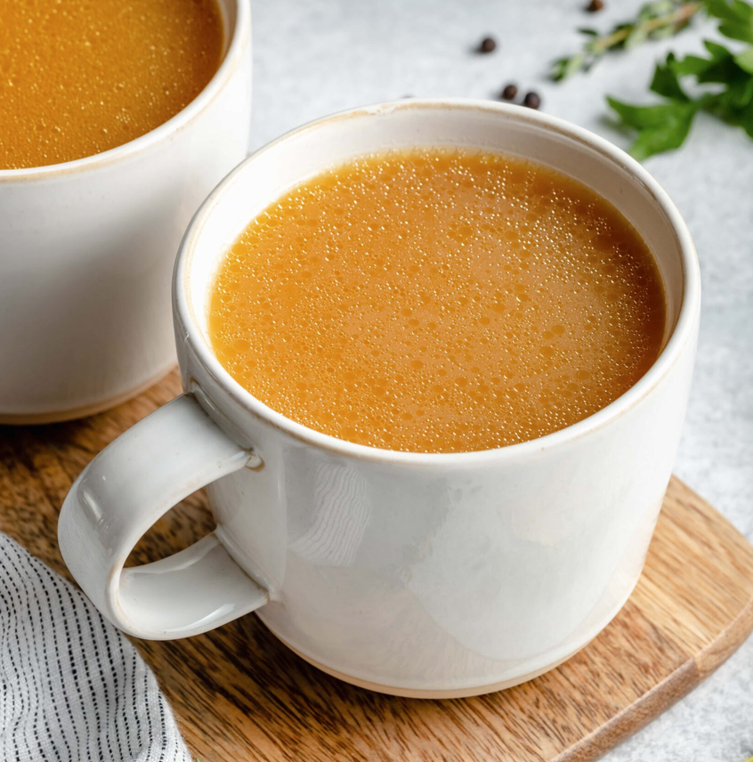 Two white mugs of golden bone broth on a wooden board