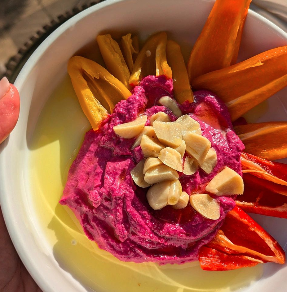 Bright pink beet hummus in a white bowl with sliced peppers and pickled garlic