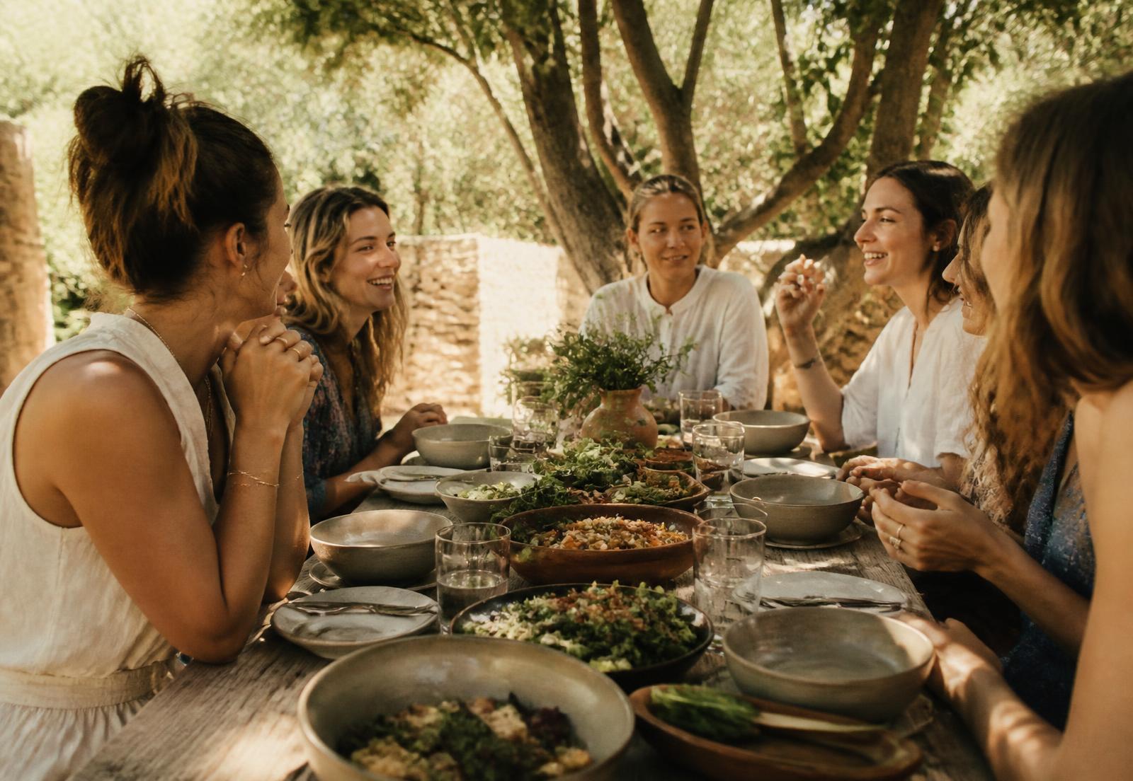 Women in conversation around a long shared table set with whole foods, outdoors under trees