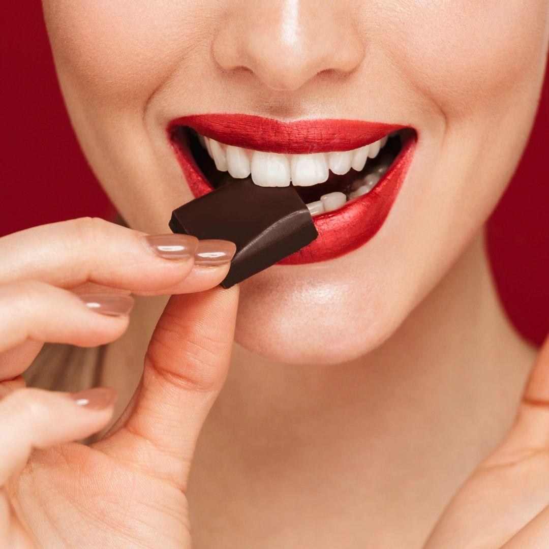 A close-up of a woman with red lipstick eating a square of dark chocolate