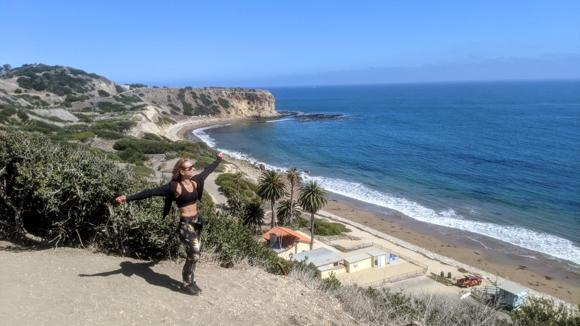 Violetta Markelou standing with arms outstretched on a coastal overlook with the ocean behind her